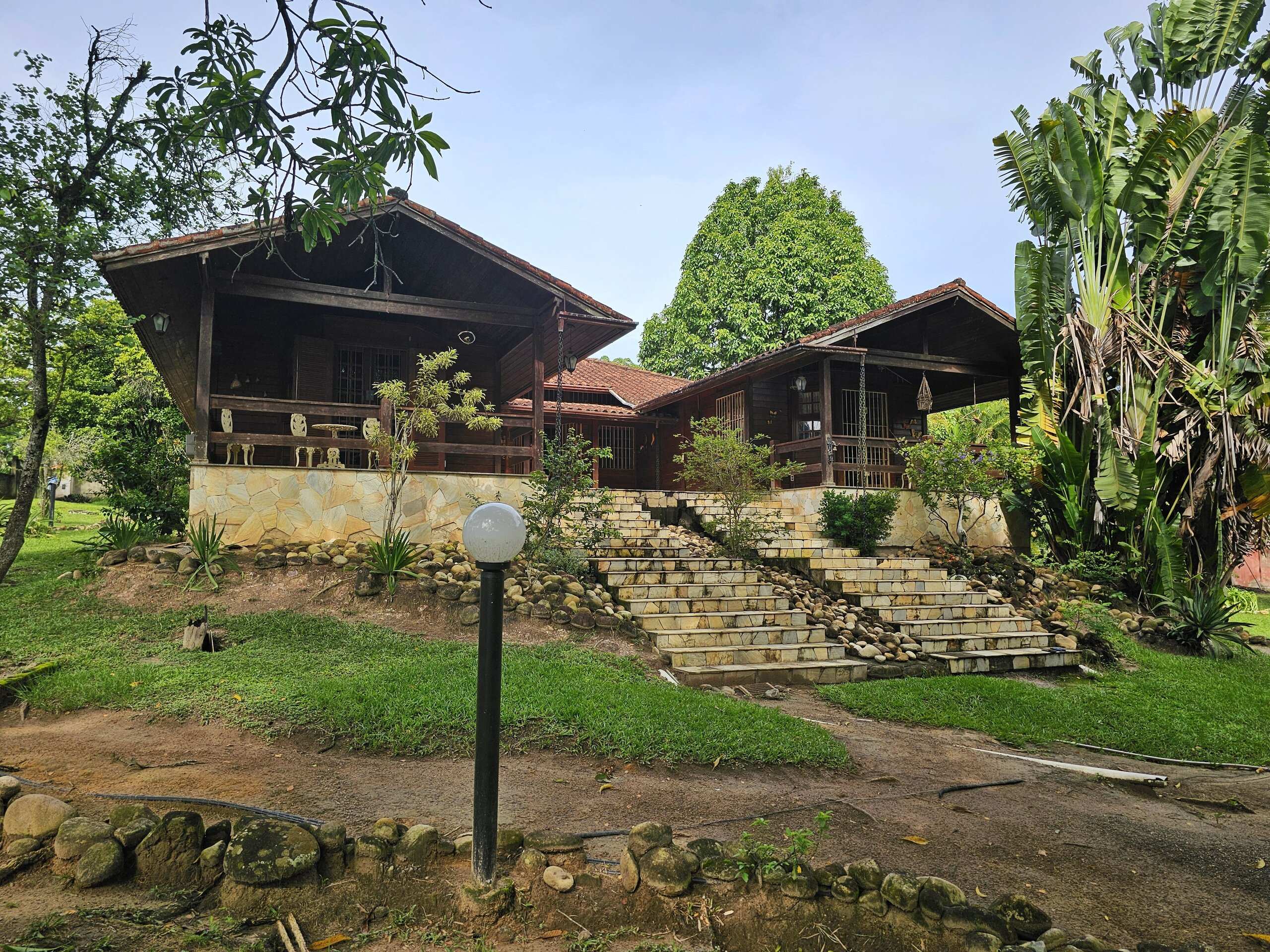Fachada da casa-mãe: chalé de madeira com escadaria de pedra subindo entre o jardim arborizado, sob céu azul