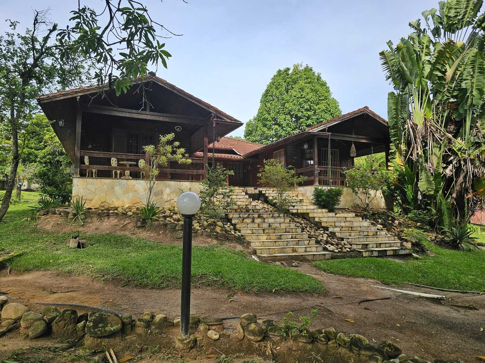 Fachada da casa-mãe com escadaria de pedra em leque, poste de jardim e cabanas de madeira sob o verde