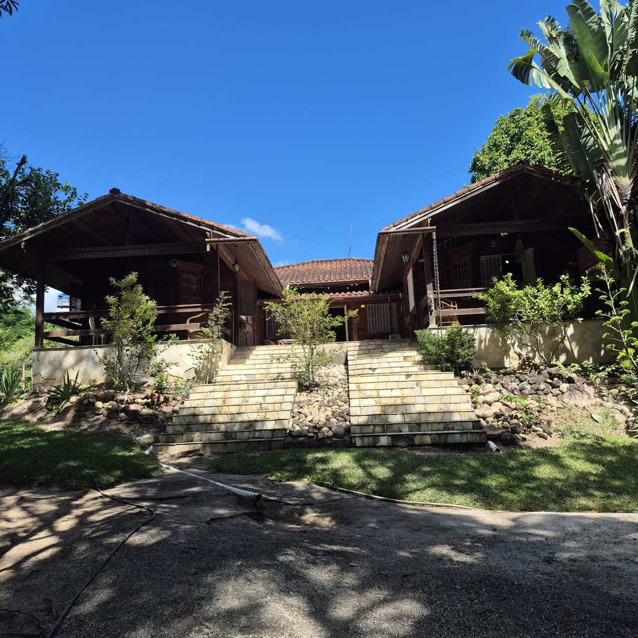 Fachada frontal das duas cabanas de madeira geminadas da casa-mãe, com escadaria de pedra, sob céu azul