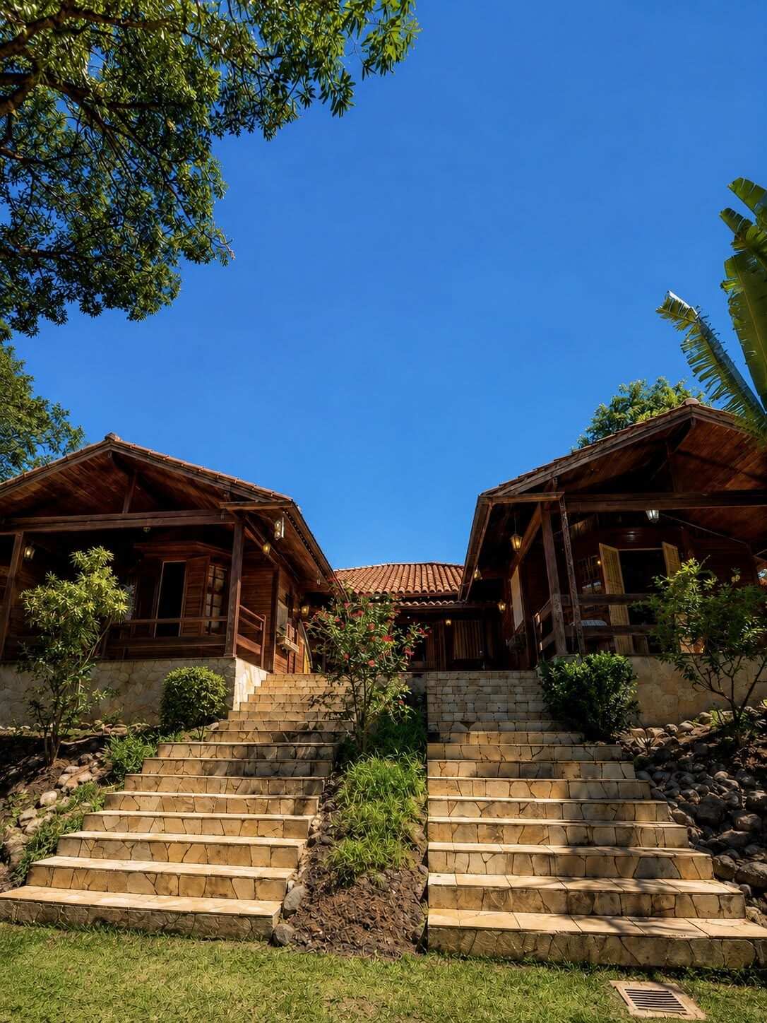 Fachada da casa-mãe: duas cabanas de madeira geminadas com escadaria dupla de pedra em leque subindo pela grama, sob céu azul limpo