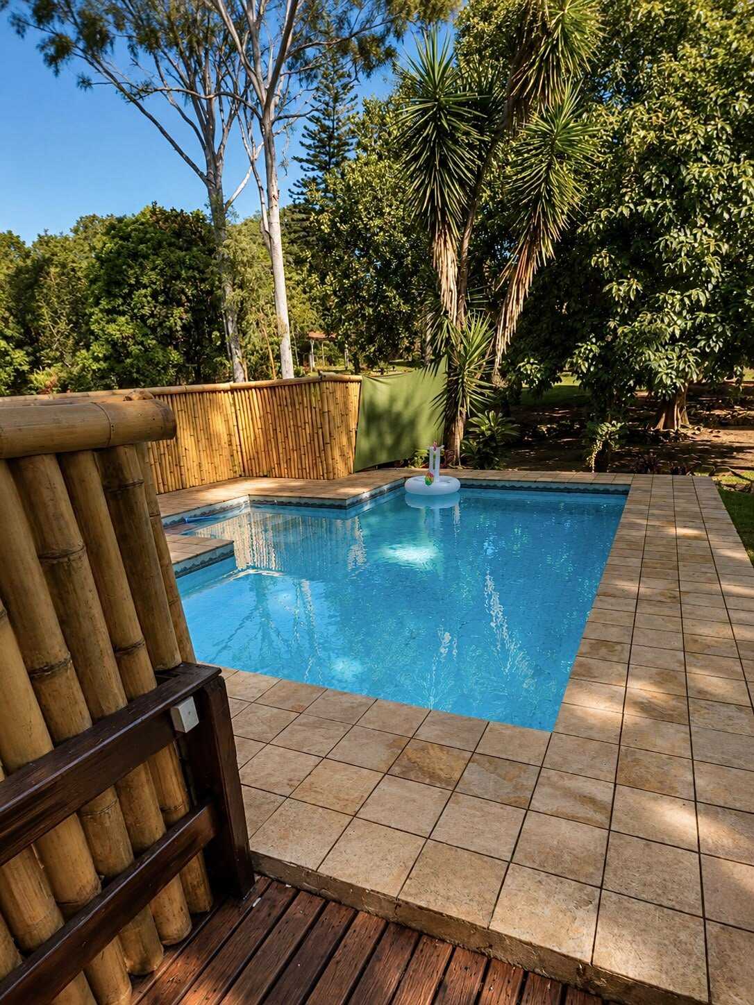 Piscina de água azul com boia flutuante, cercada por deck de madeira, muro de bambu e palmeiras