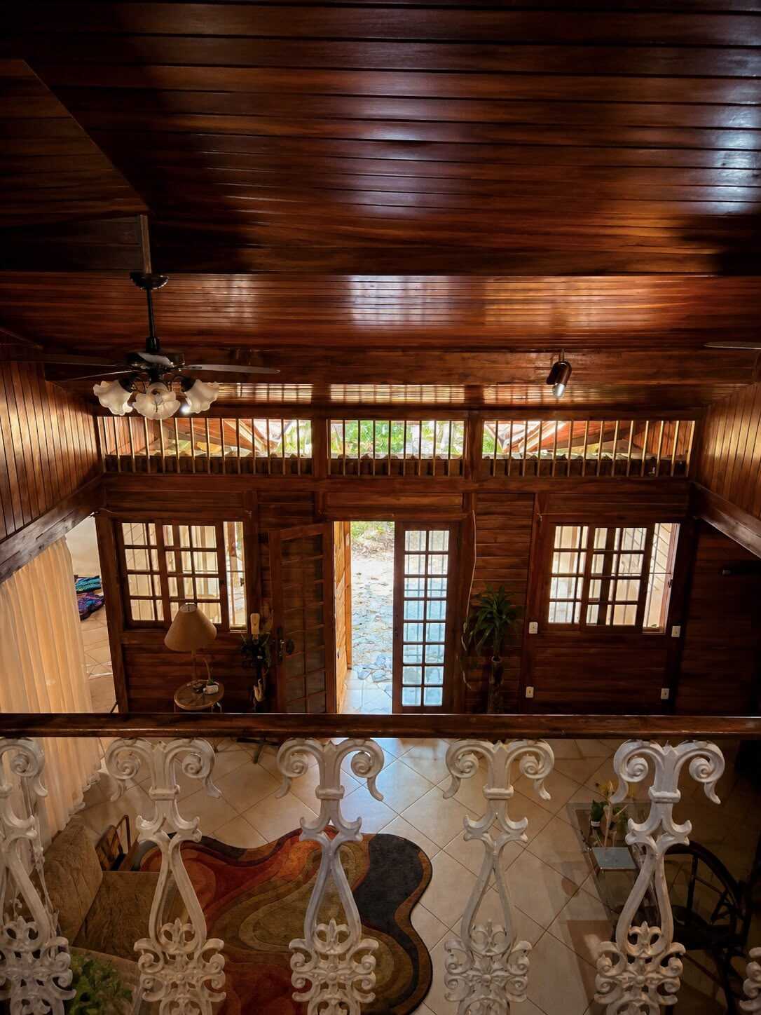 Interior de madeira envernizada da casa-mãe visto do mezanino, com portas de vidro ao fundo e guarda-corpo branco trabalhado em primeiro plano